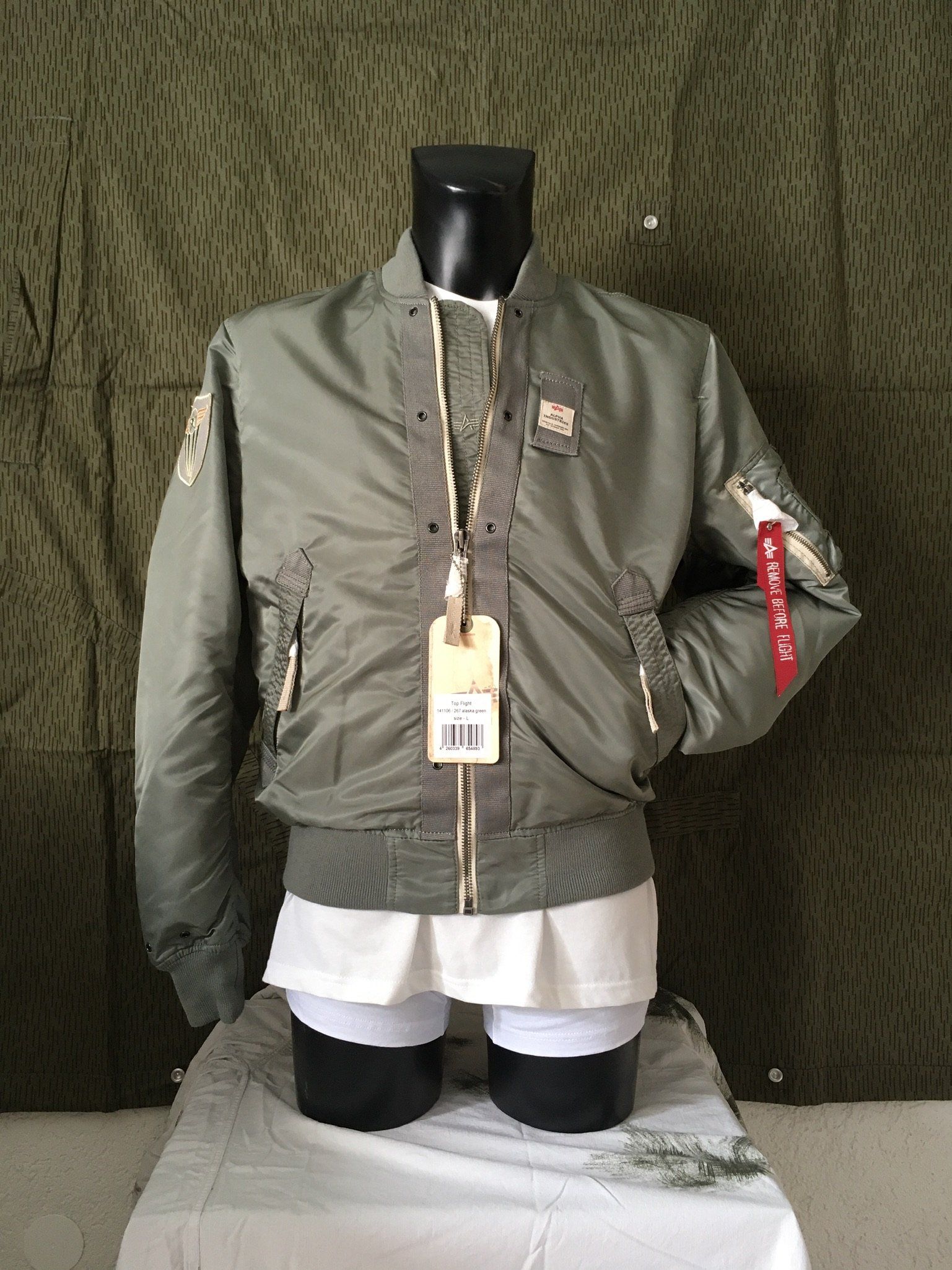 Grüne Bomberjacke an einer schwarzen Schaufensterpuppe, mit weißem Hemd und Preisschild, vor einem grünen Tarnhintergrund.