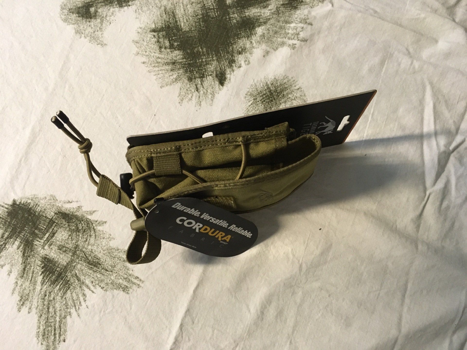 Olivgrüne Cordura-Gebrauchstasche mit Kordelzug und Etikett, auf einem gemusterten Tuch liegend.