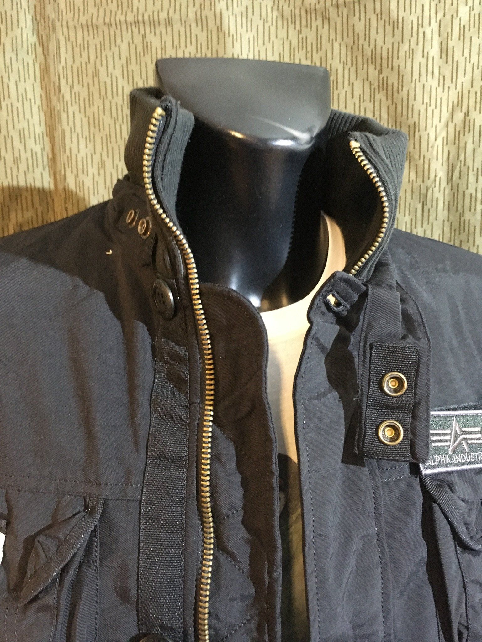 Schwarze Jacke an einer Schaufensterpuppe mit hohem Kragen und Reißverschluss, mit Taschen und dem Alpha Industries-Logo.