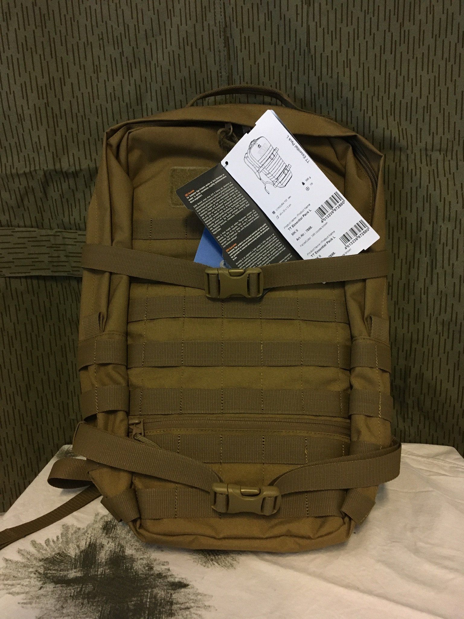 Hellbrauner taktischer Rucksack mit MOLLE-Gurtband und Schnallen, auf einer weißen Oberfläche liegend, Etikett sichtbar.