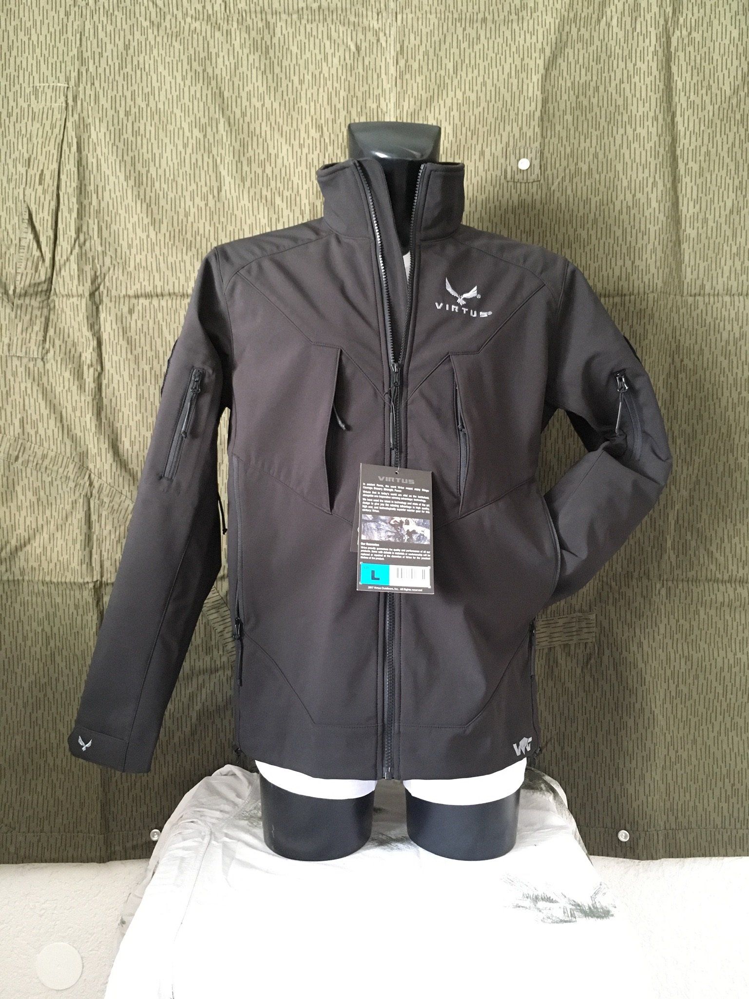 Schwarze Jacke an einer Schaufensterpuppe, mit hohem Kragen und mehreren Taschen, vor einem Tarnhintergrund.