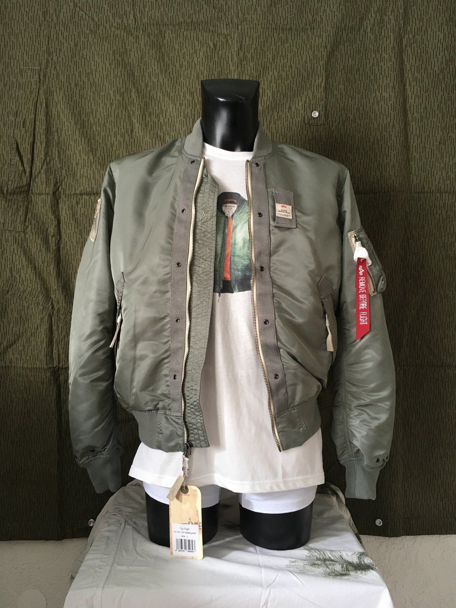 Grüne Bomberjacke auf einer Schaufensterpuppe, mit weißem T-Shirt darunter, vor gemustertem grünen Hintergrund stehend.