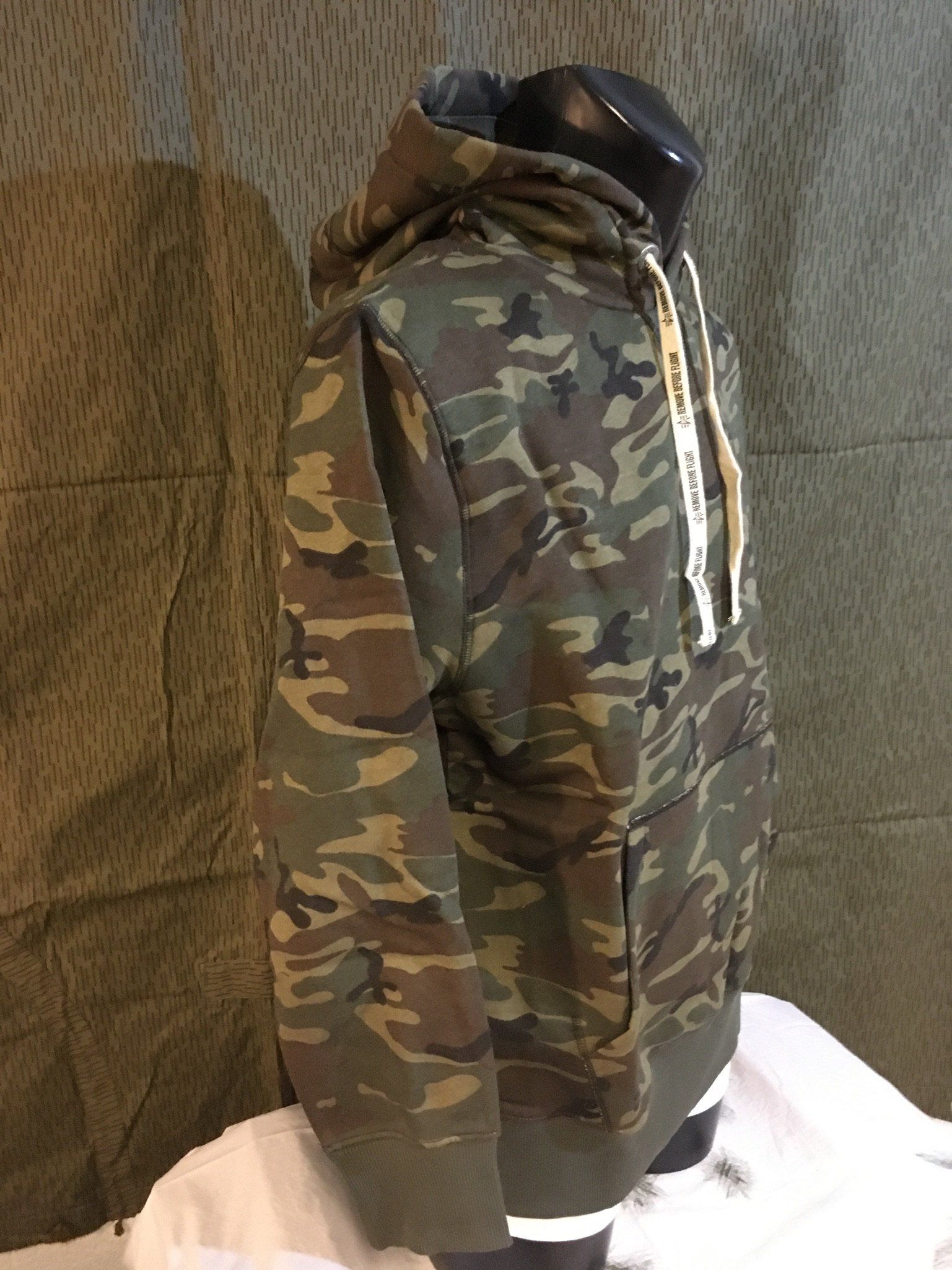 Camouflage-Kapuzenpullover auf einer Schaufensterpuppe, mit weißen Kordeln, vor grünem Hintergrund.