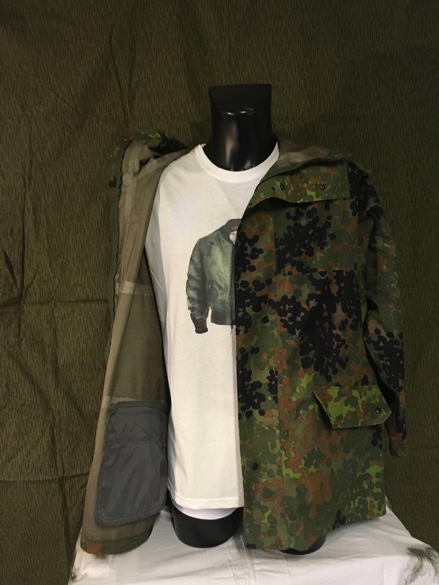 Schaufensterpuppe mit weißem T-Shirt, offener Tarnjacke und offener olivfarbener Jacke vor einem Tarnhintergrund.