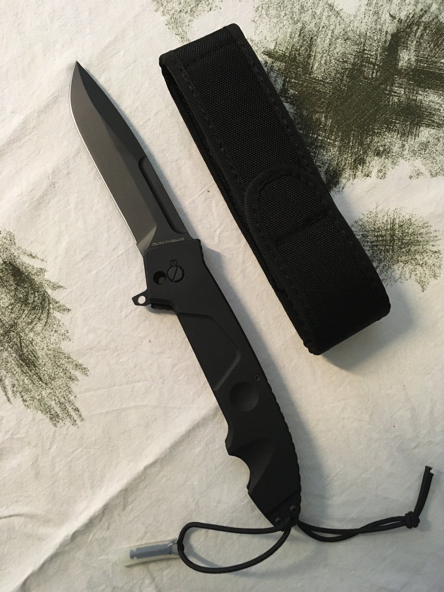 Schwarzes taktisches Messer mit gezackter Klinge und Scheide auf gemustertem Stoff.