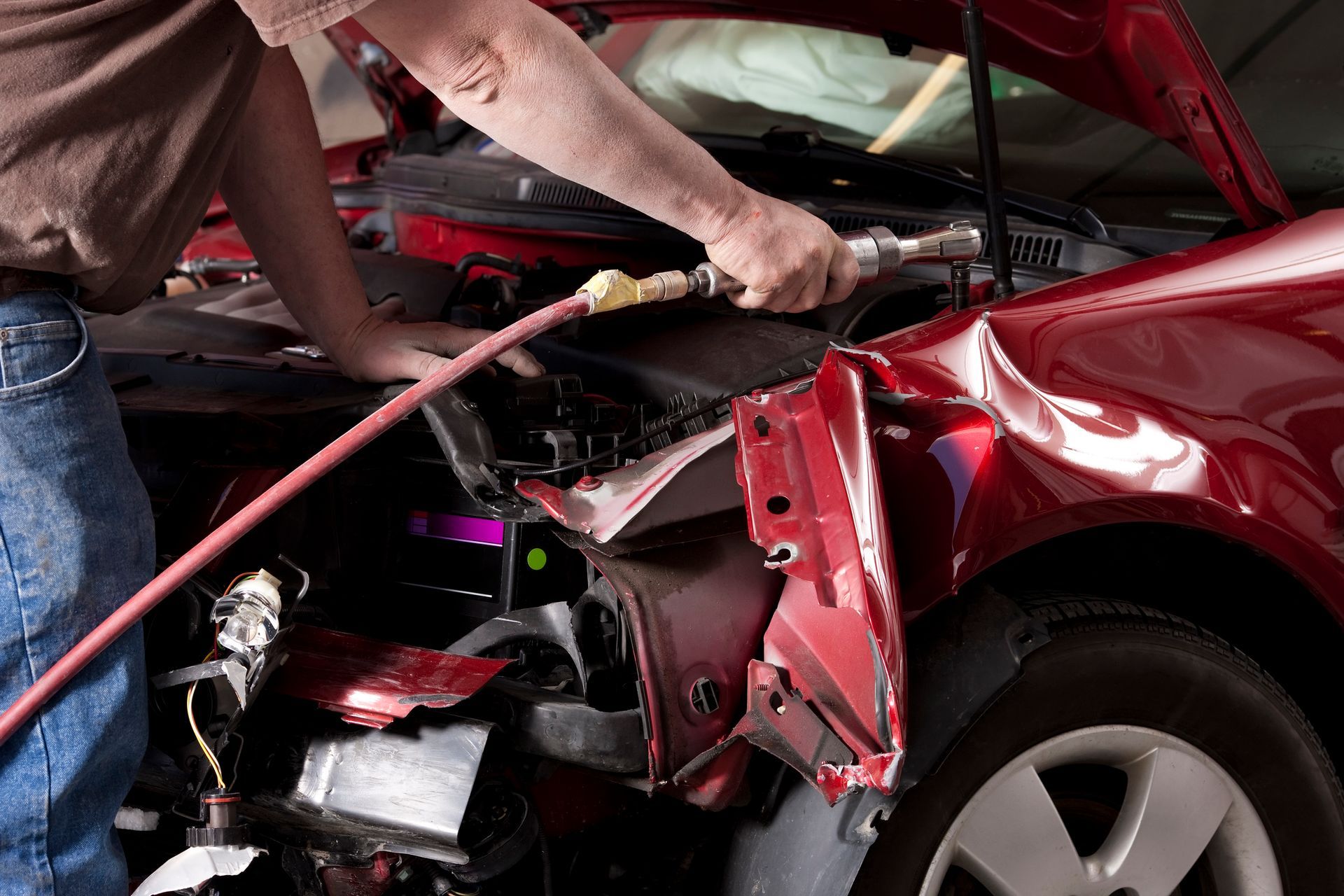 A man repairs a red car using a tool with a hose, showcasing hands-on work at a car body shop.