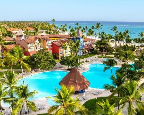 Aerial view of a tropical resort with a large blue swimming pool, palm trees, and hotel buildings near a sandy beach.