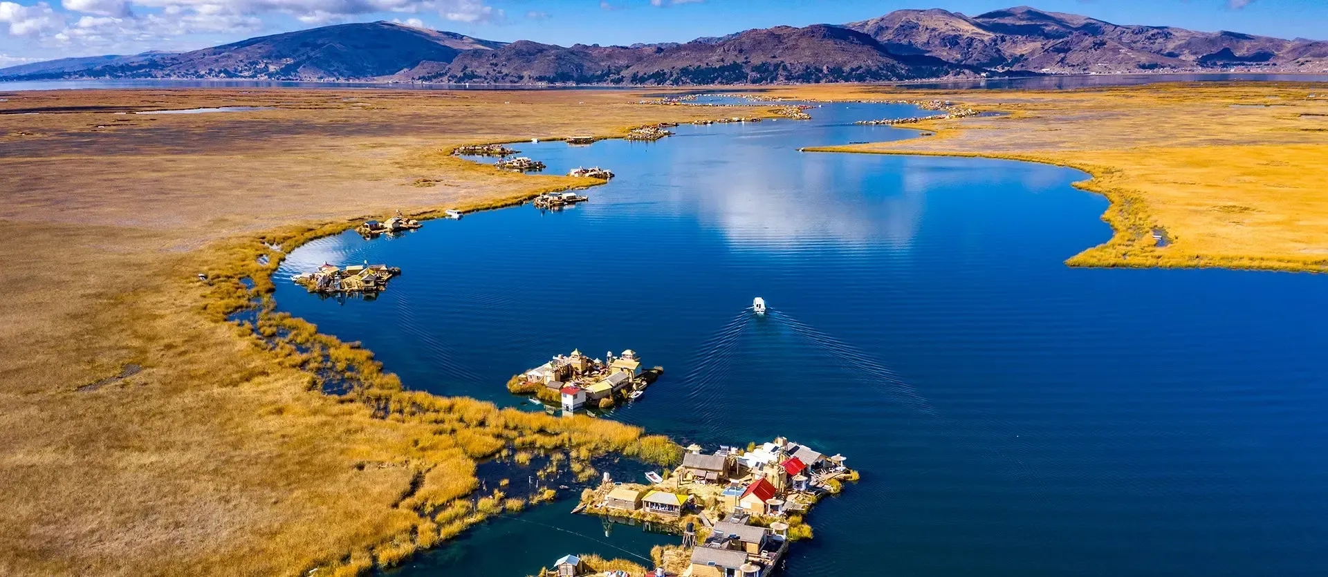 Aerial view of a blue lake beside golden grasslands, with small boats and mountains in the distance