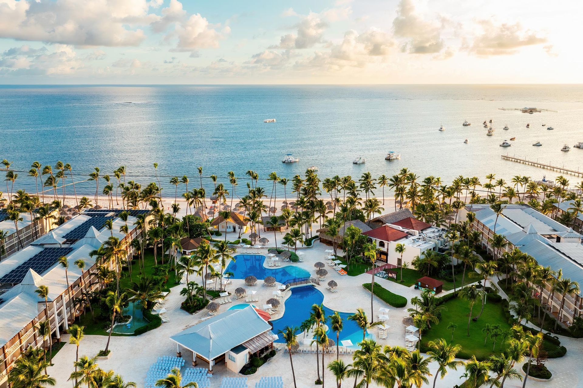 Aerial view of a tropical resort with a blue pool, palm trees, and white buildings along a sunny beach with the ocean.