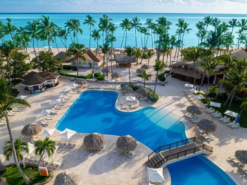 An aerial view of a tropical resort with a turquoise swimming pool, thatched-roof cabanas, palm trees, and a sandy beach.