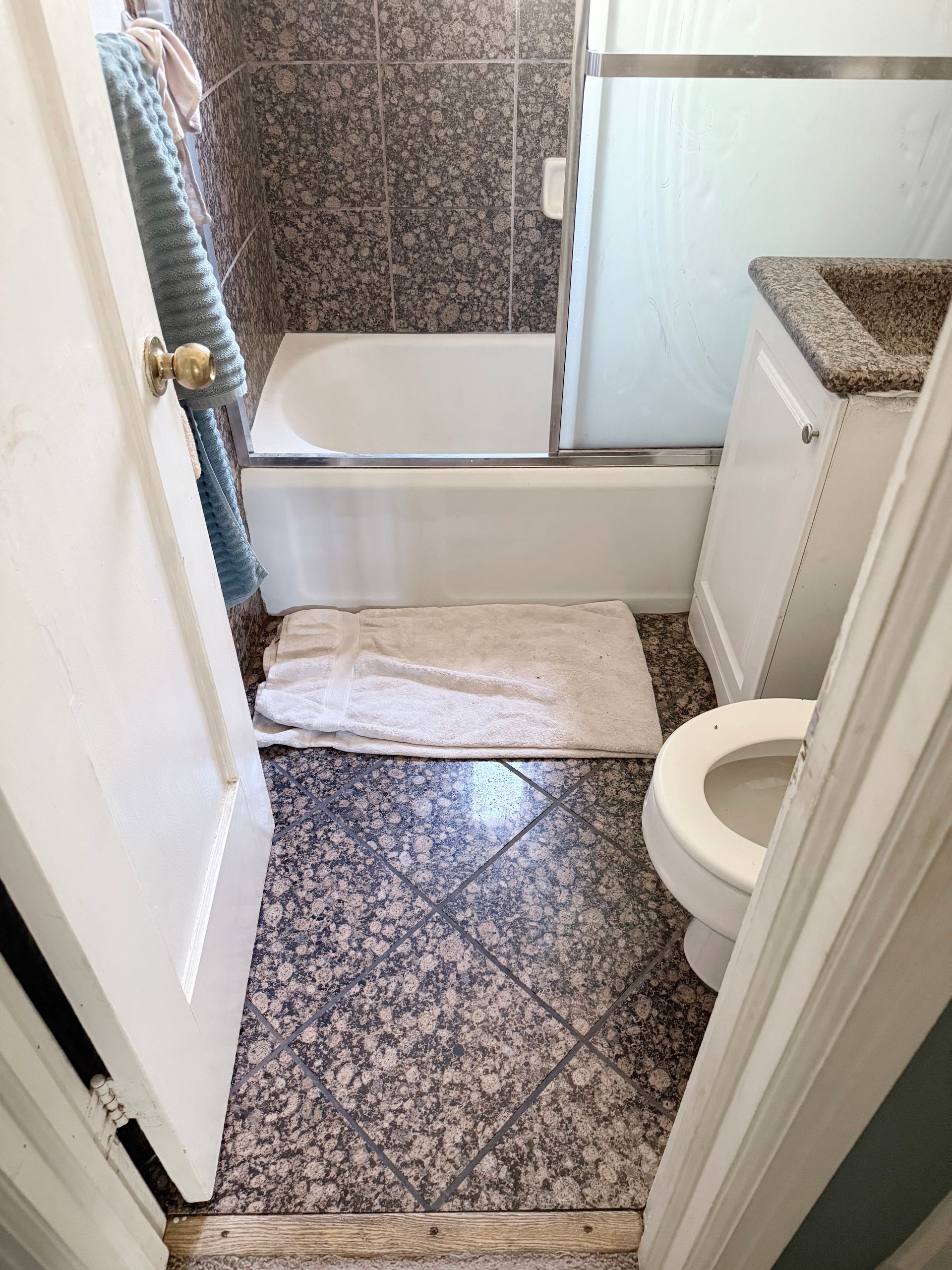 Small bathroom with granite tile floor and walls, a white tub, toilet, and sink, and a partially open door.
