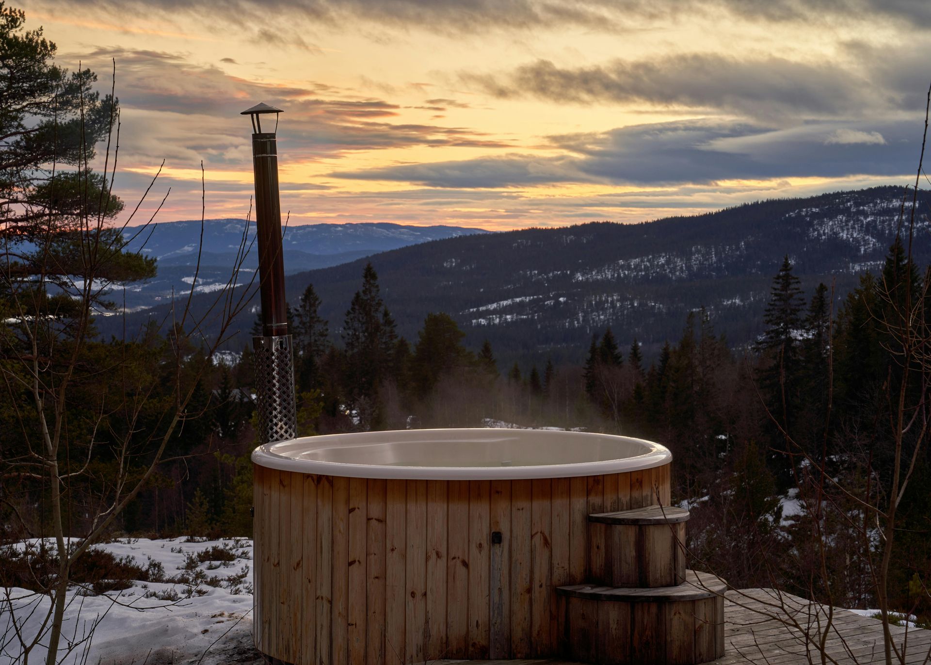 Wooden hot tub with steam, overlooking a snowy mountain landscape at sunset.