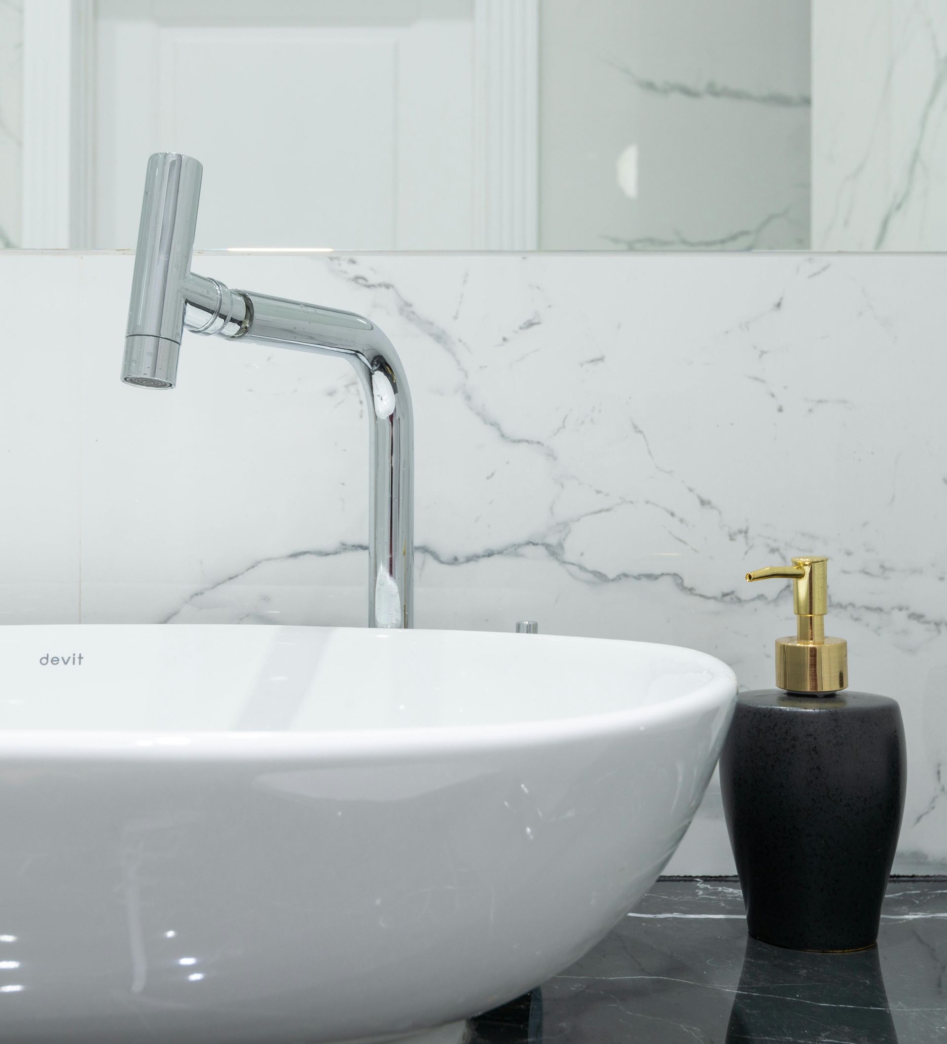 White sink with chrome faucet and black soap dispenser with gold pump against marble wall.