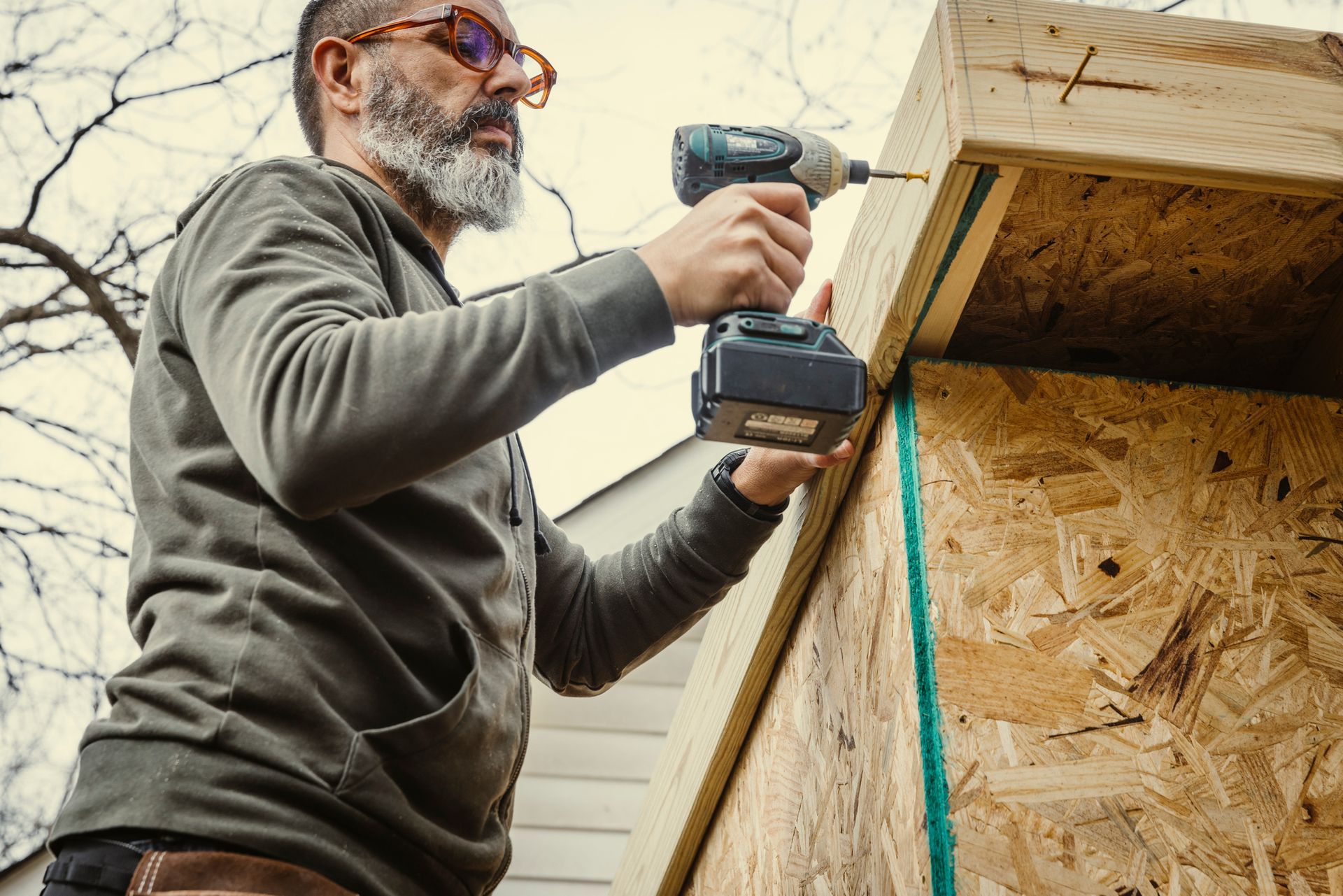 Man with beard using a power drill to assemble a wooden structure outdoors.