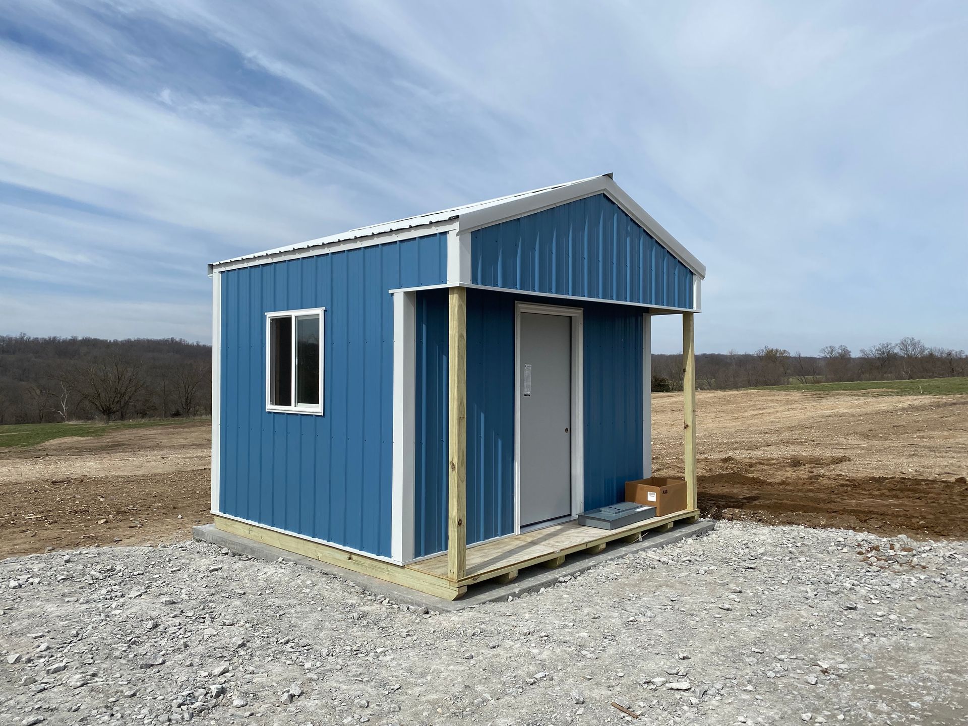 Blue metal shed with a white roof, window, and door, set on gravel, under a blue sky.