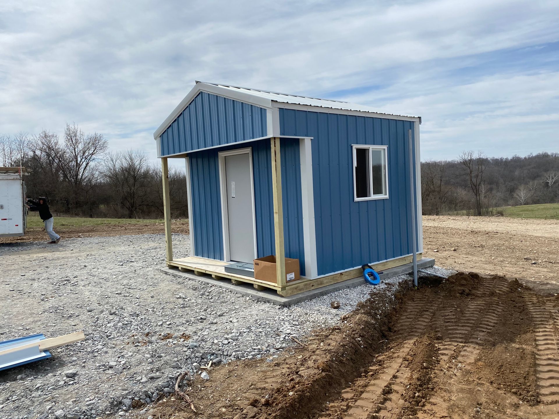 Blue shed with porch under construction, gravel ground, person nearby.