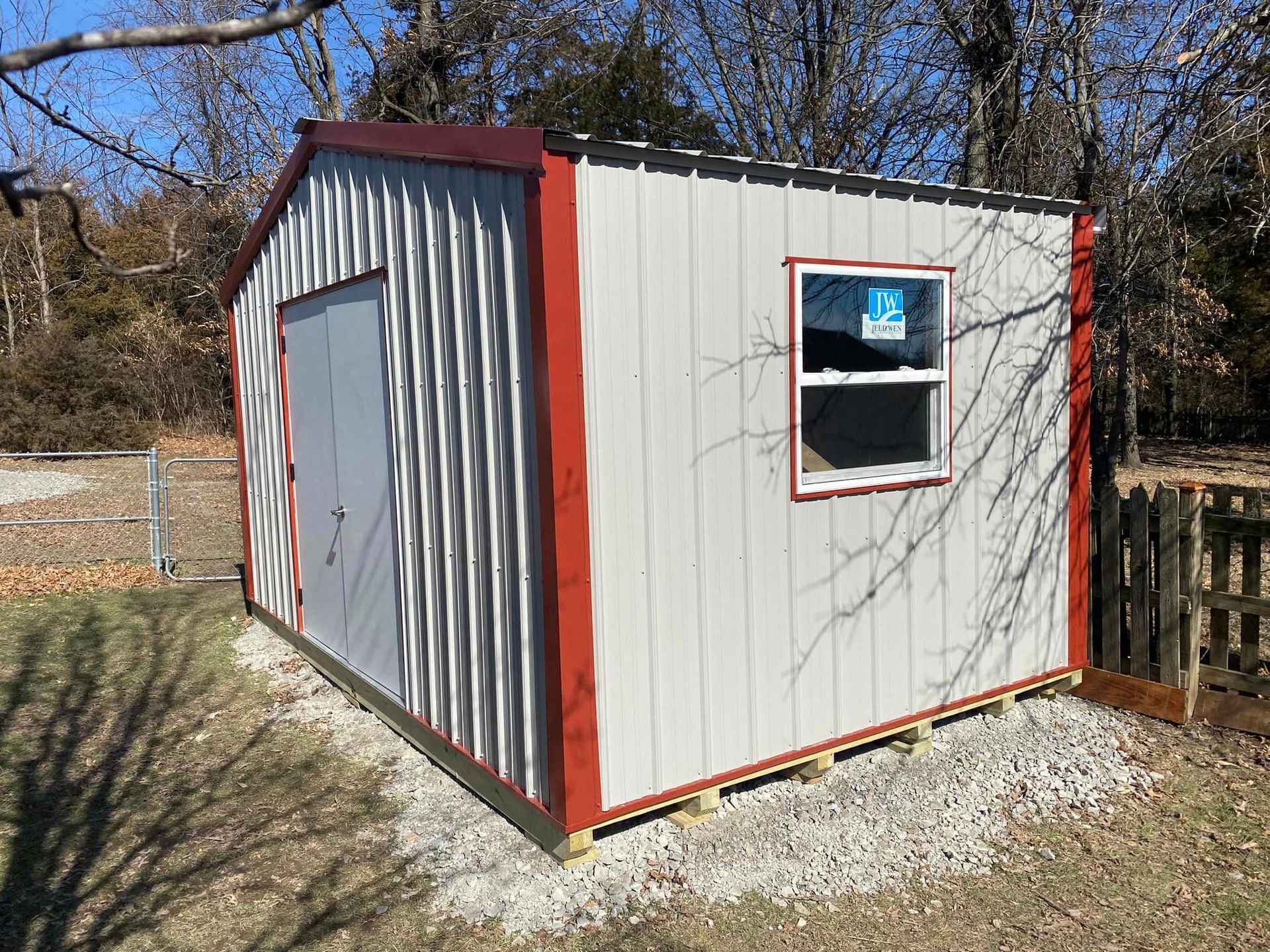 Metal shed with gray door and window, red trim, on gravel in a yard.