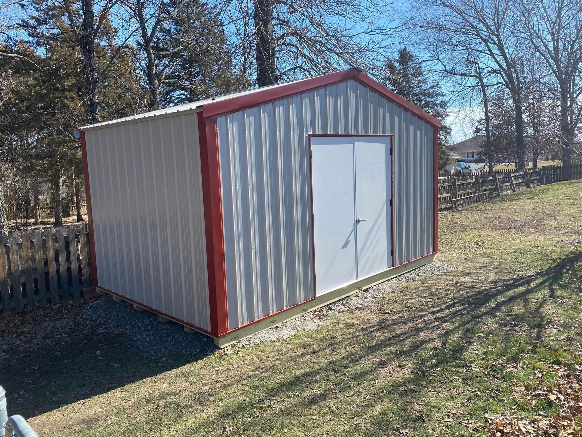 Metal storage shed with red trim and double white doors, sitting on a grassy hill with a wooden fence in the background.