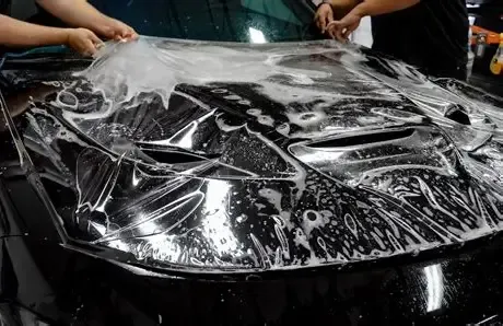 Person wrapping a black car hood with clear protective film in a workshop