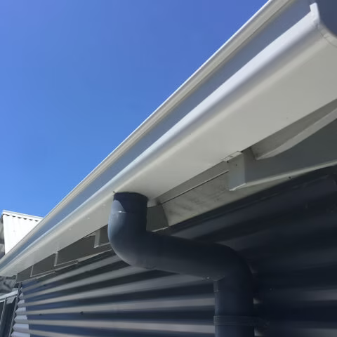 White gutter with a downspout attached to a corrugated metal building under a clear blue sky.— Gutter Guard In Laurieton, NSW