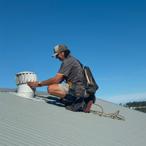 Roofer installing a ventilation fan on a metal roof, wearing safety gear, under a blue sky.— Gutter Guard In Lake Cathie, NSW