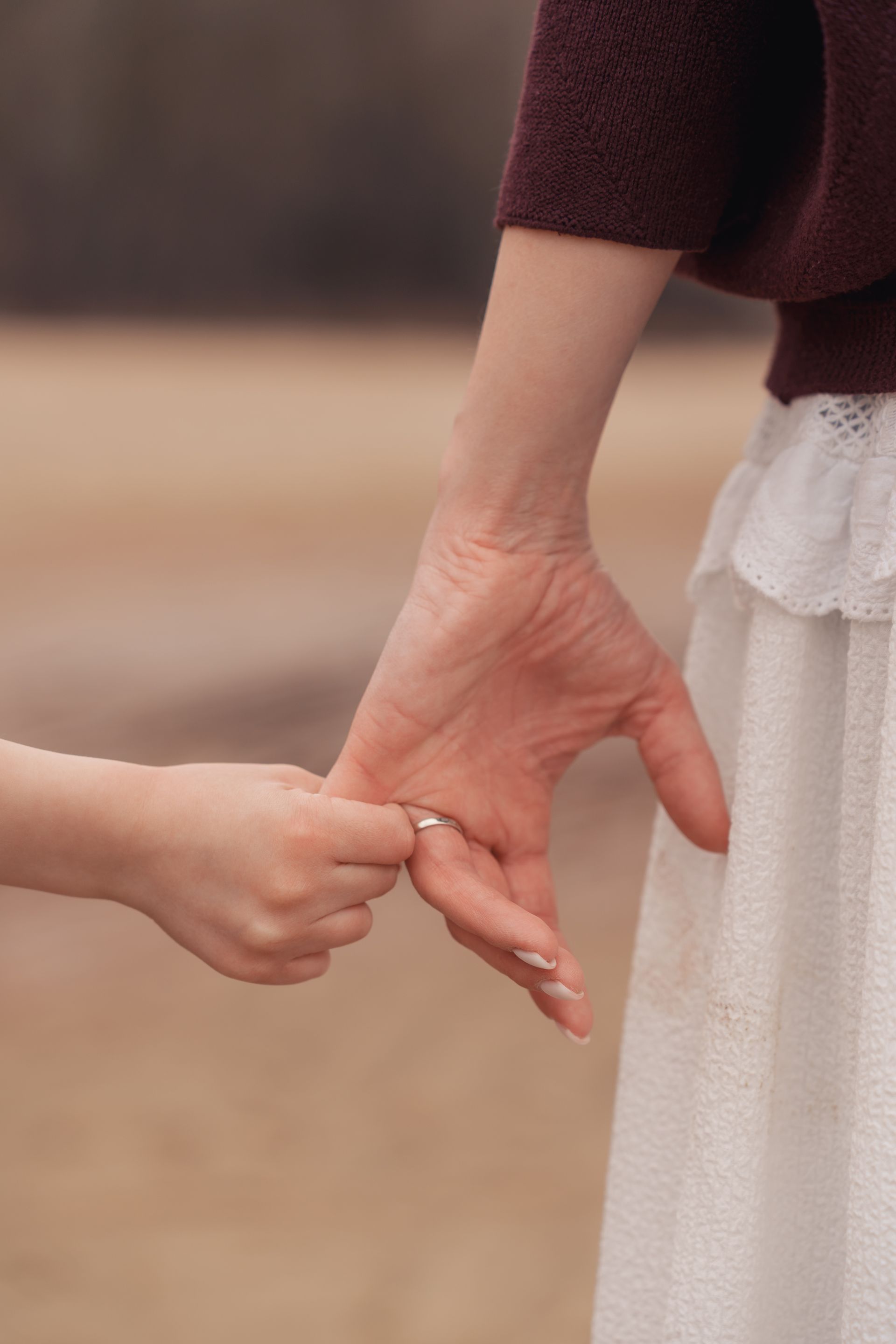 Two hands gently holding, with a maroon sleeve and white dress visible against a blurred outdoor background Two hands gently holding, with a maroon sleeve and white dress visible against a blurred outdoor background