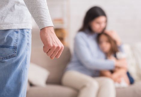 A person stands in foreground while a woman hugs a child on a couch in the background A person stands in foreground while a woman hugs a child on a couch in the background