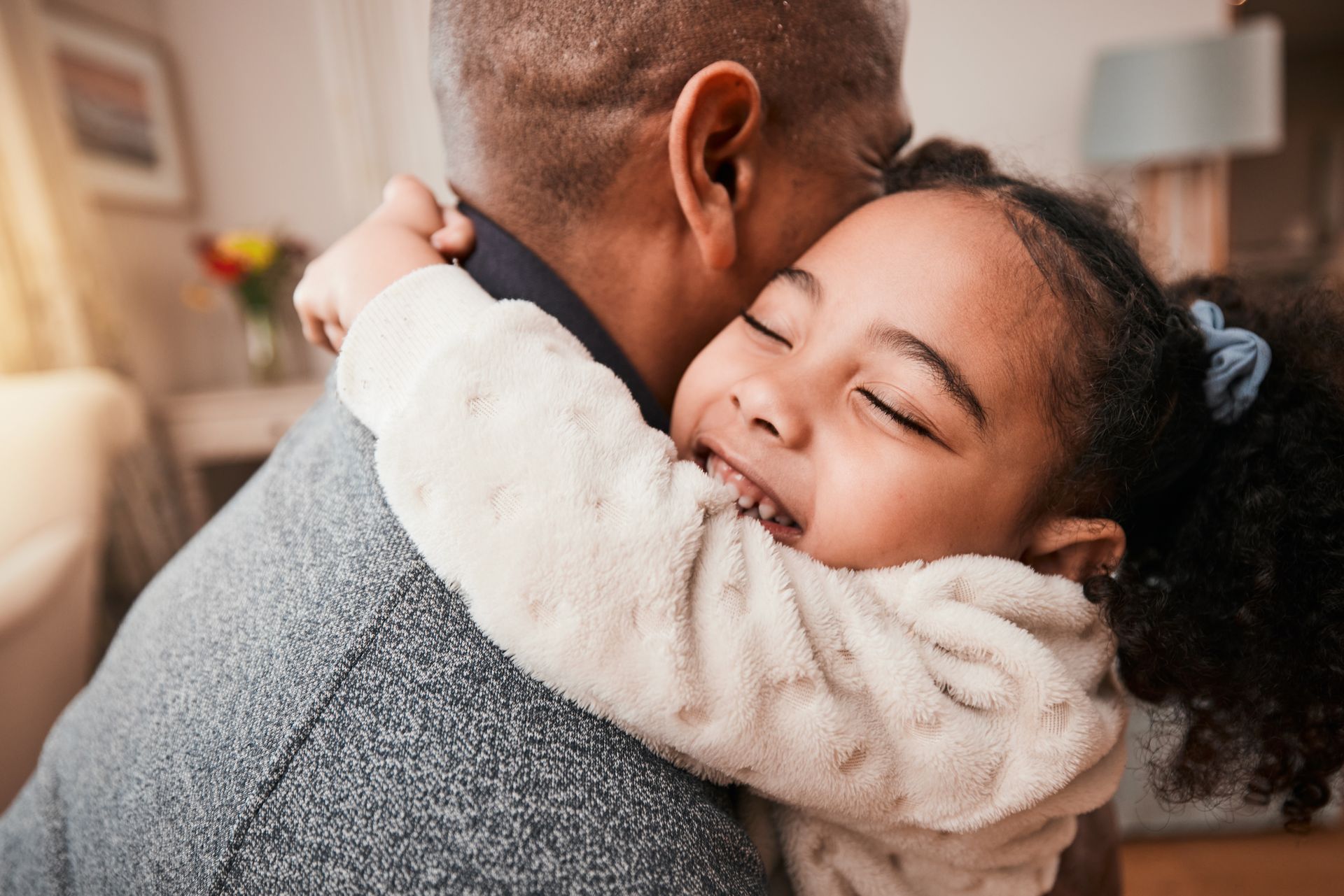 Young child hugging an adult indoors, both smiling warmly in a cozy living room. Young child hugging an adult indoors, both smiling warmly in a cozy living room.