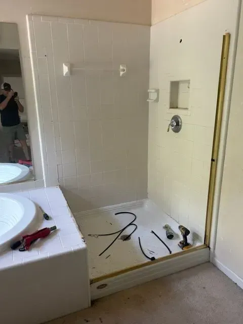 Bathroom with a shower stall under renovation, showing exposed plumbing and partially dismantled fixtures.