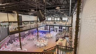 Spacious event venue with tables, white chairs, two levels, and floral decorations.