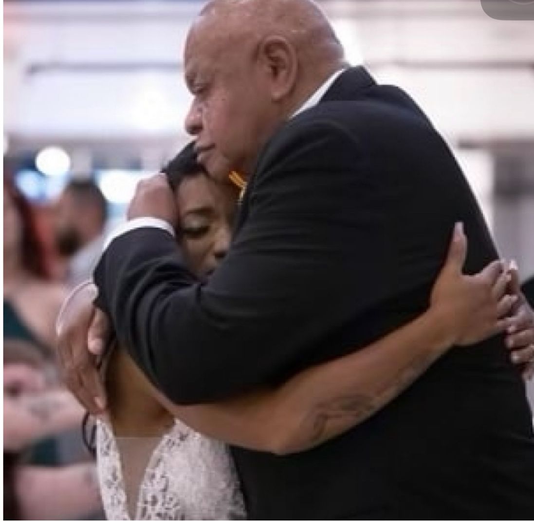 Man in suit embraces person in white dress, likely a wedding.