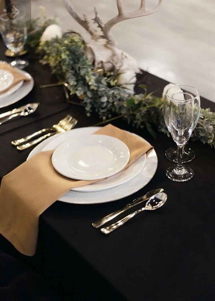 Formal dining table setting with white plates, gold silverware, and tan napkins on a black tablecloth, with garland and antlers in the background.