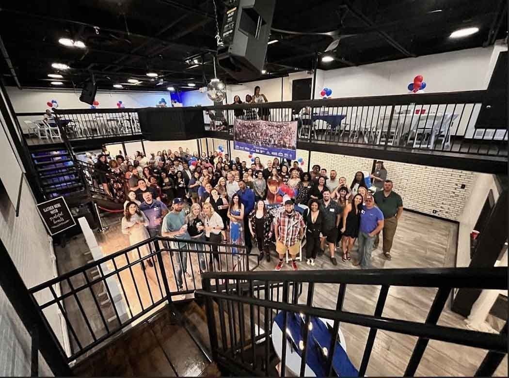 Large group of people gathered inside a gym, standing near a stage and stairs. Some are looking at the camera.