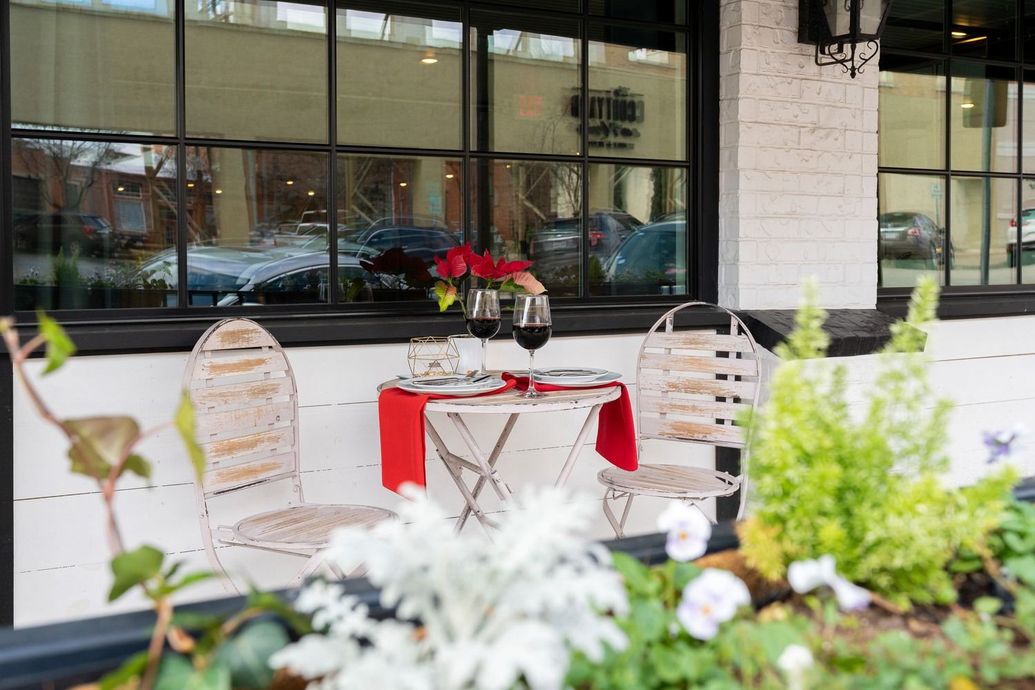 Outdoor dining table set for two with red tablecloth and wine glasses, flowers.