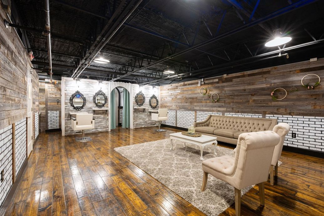 Interior of a modern salon.  Wood floors and walls.  Seating area with couch, chairs, rug, coffee table.  Mirrors, stations in the background.