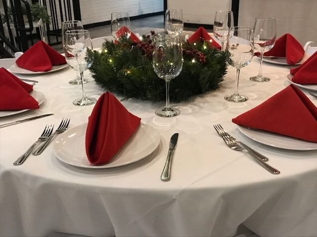Round table set for a holiday dinner with red napkins, white plates, silverware, and a wreath centerpiece.