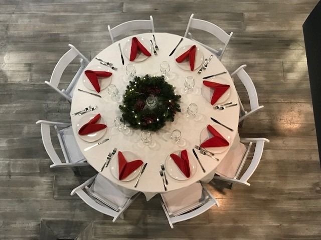 Round table set for a holiday meal, with red napkins, white chairs, and a central wreath.