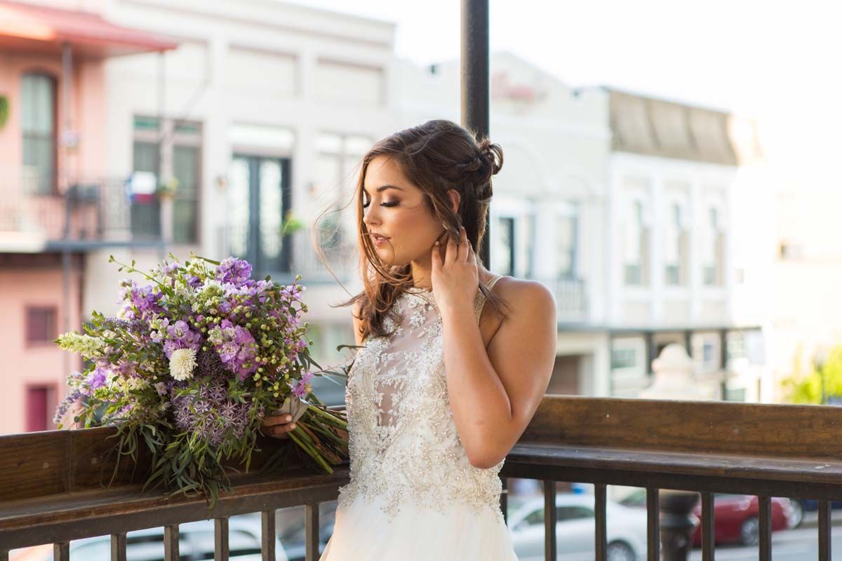 Bride with purple bouquet on a balcony, looking down. City buildings in the background.