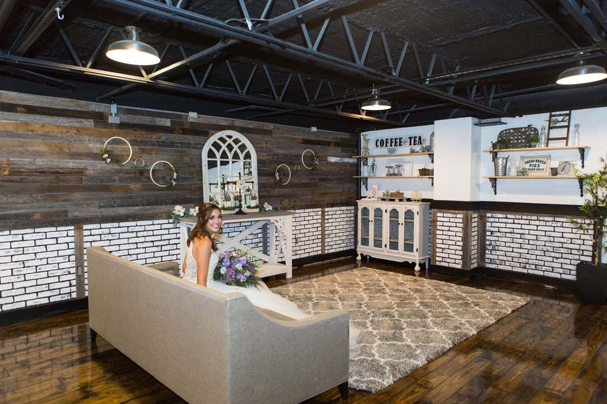 Bride sitting on a beige couch in a rustic room, holding a bouquet, with wood and brick walls.