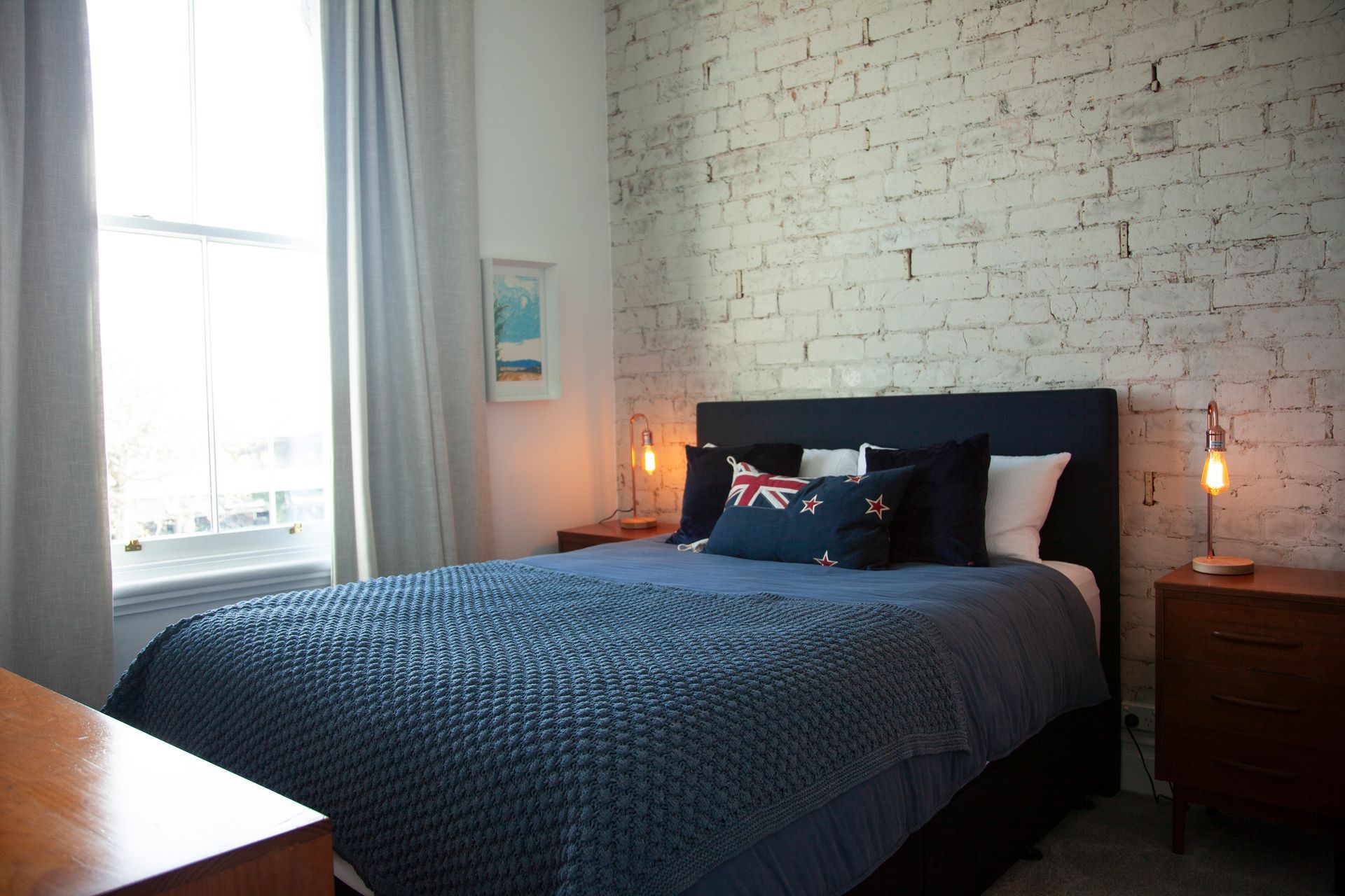 Bedroom with blue bedspread, white brick wall, window with gray curtains, and wooden nightstands.