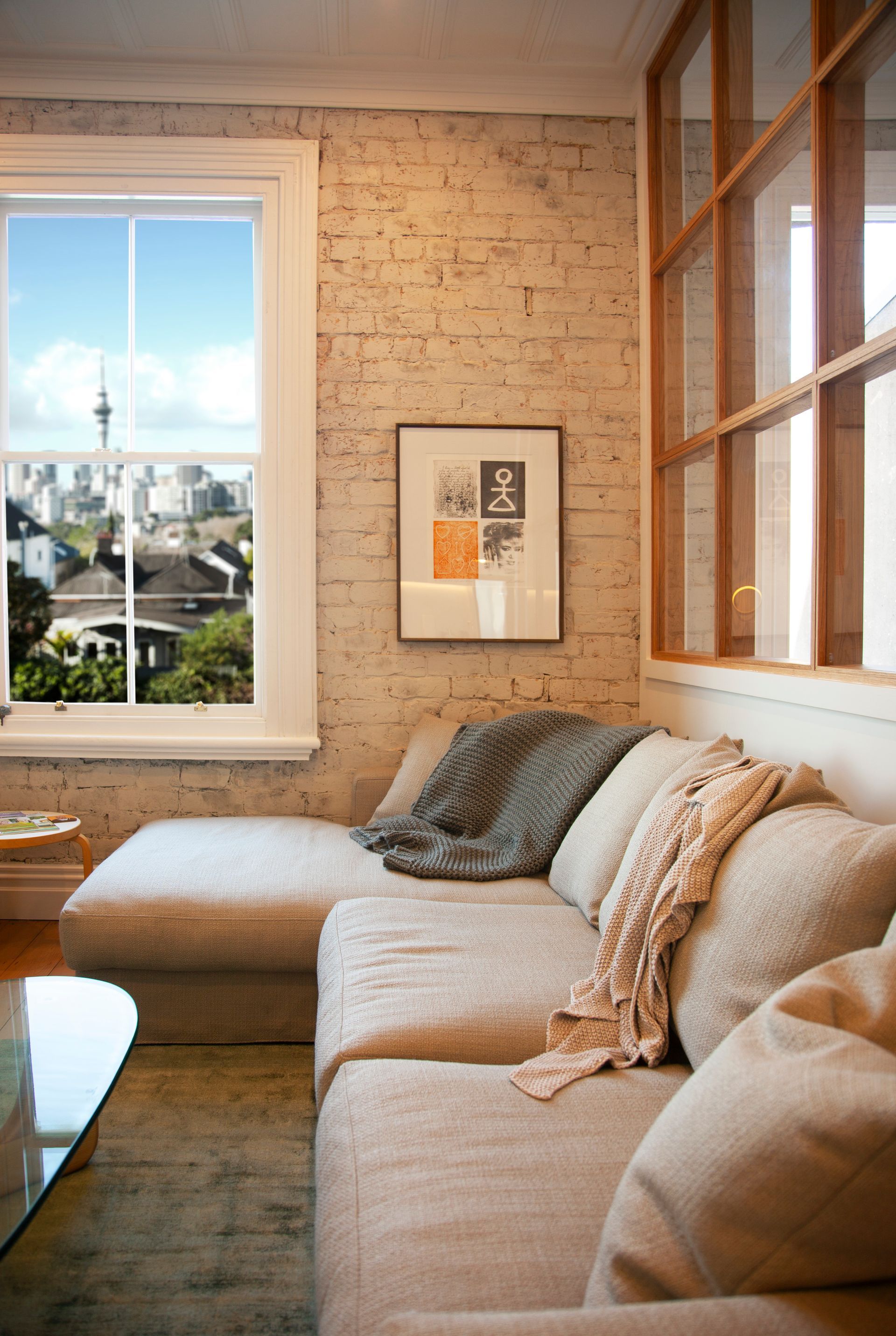 Cozy living room with a beige sectional couch, exposed brick wall, and a window with a city view.
