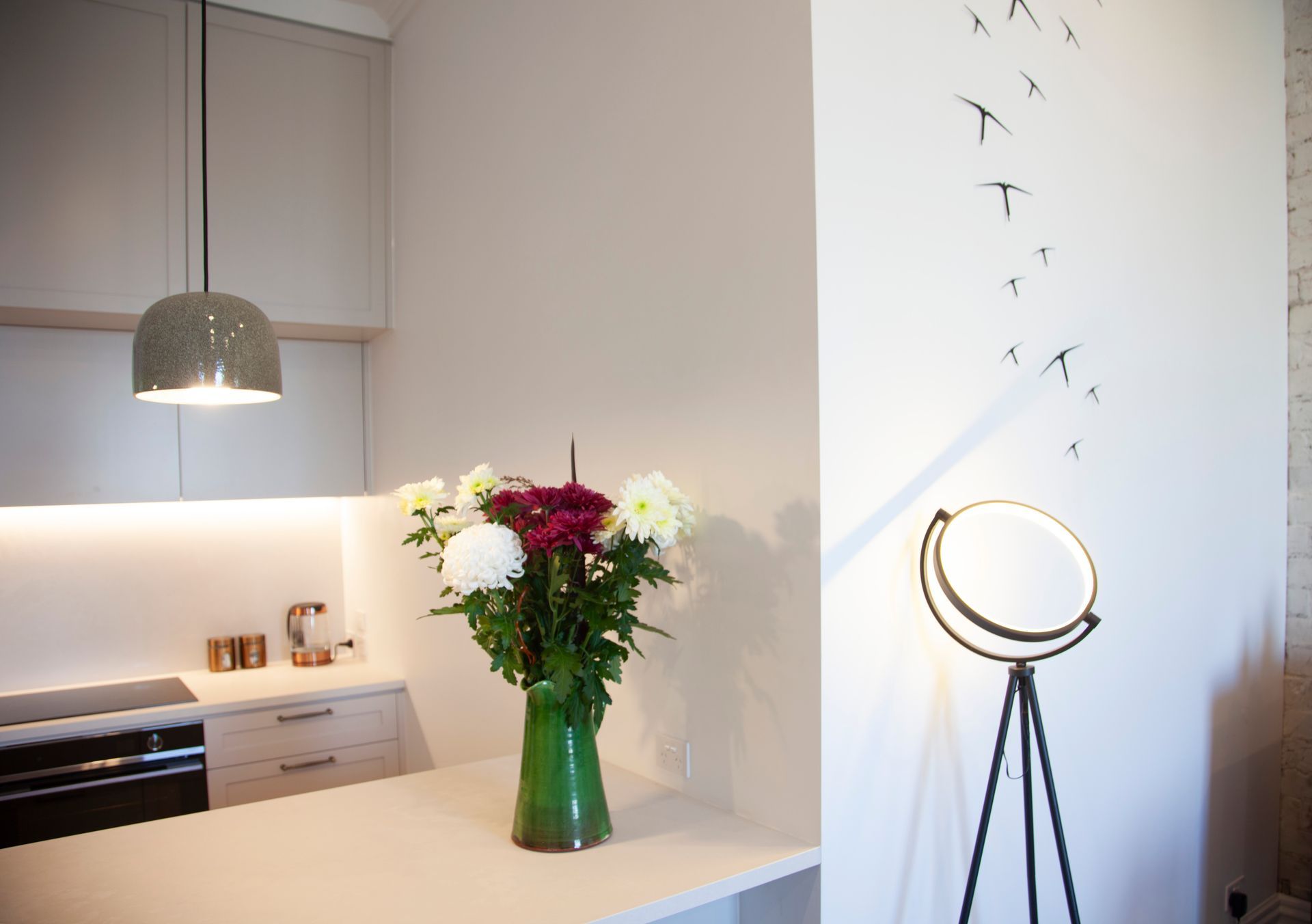 Kitchen with flowers in a vase, a hanging lamp, and a floor lamp with bird silhouettes on the wall.
