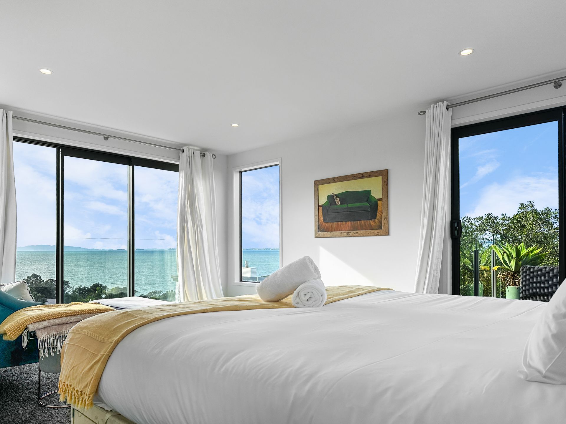 Bedroom with large windows overlooking the ocean; white bed, yellow blanket, art on wall.