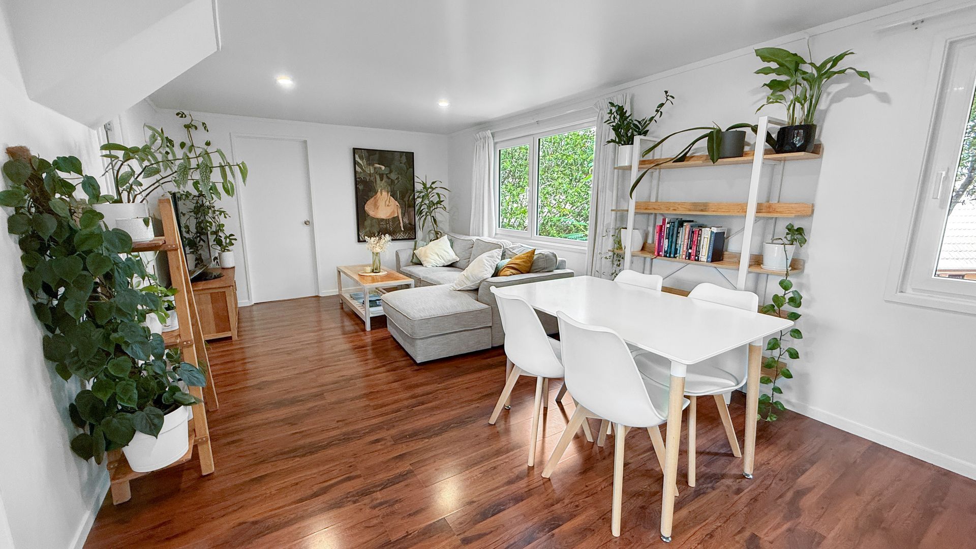 Living room with hardwood floors, white walls, a dining table, and a sofa, adorned with many plants.