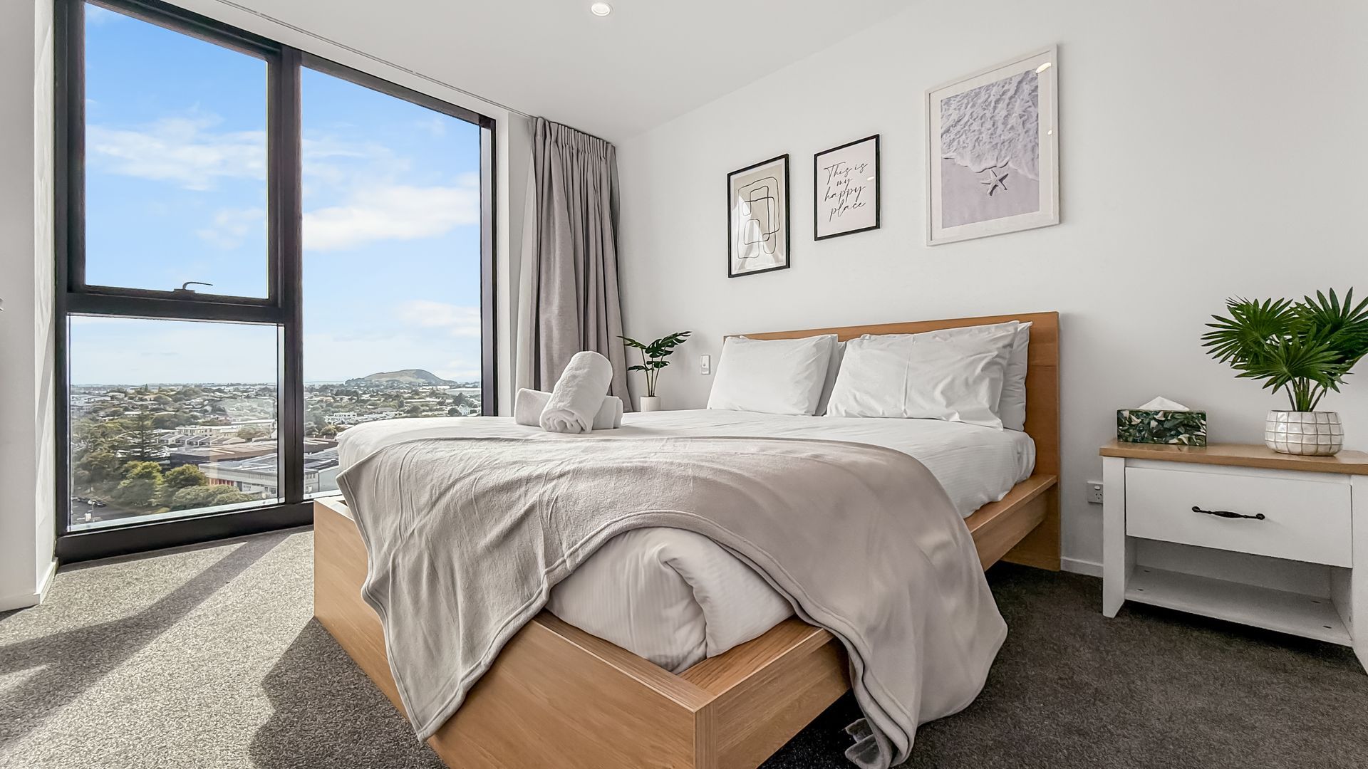 Bedroom with bed, large window with cityscape view, neutral tones, artwork on wall.