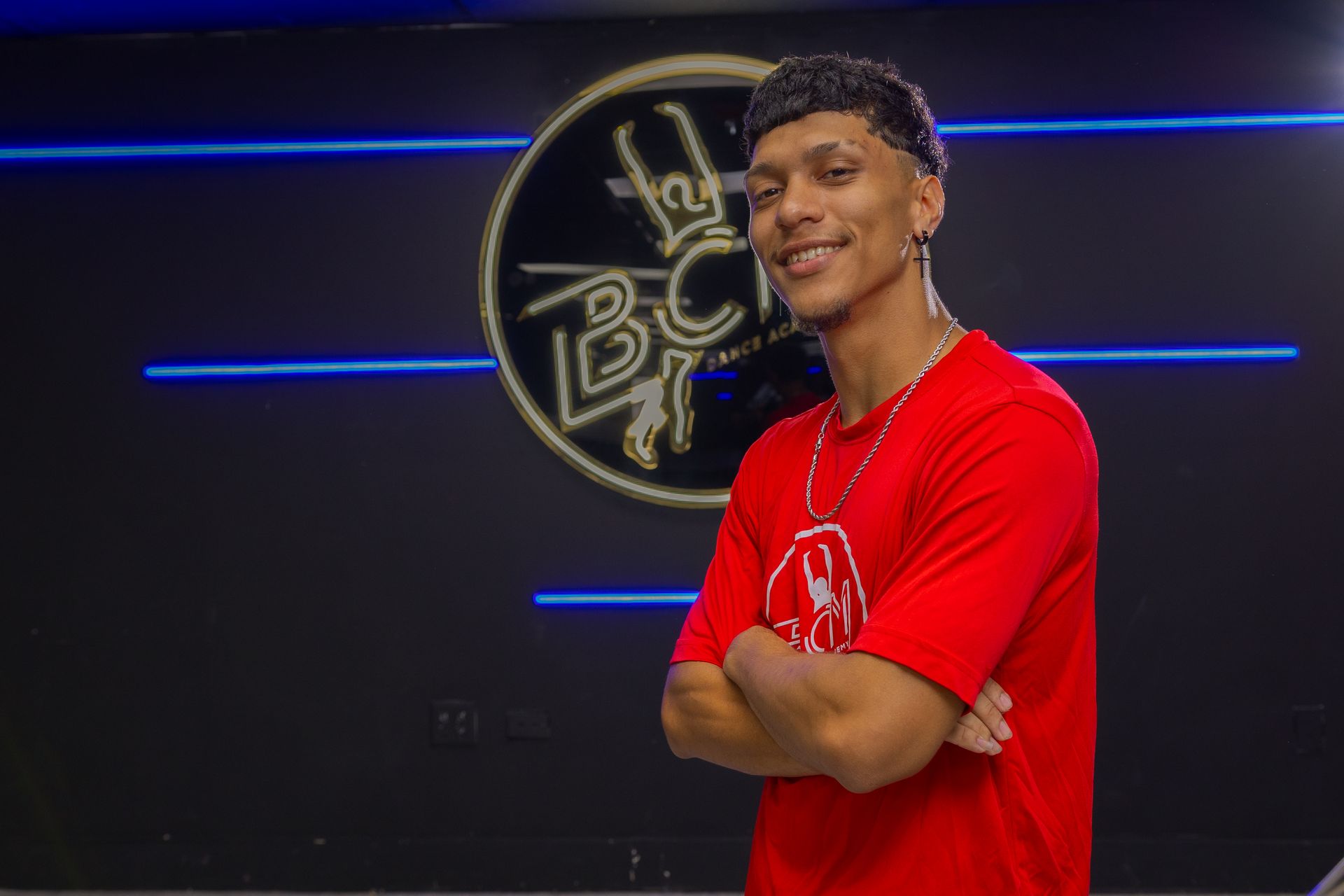 A young man in a red shirt is standing with his arms crossed in front of a sign.