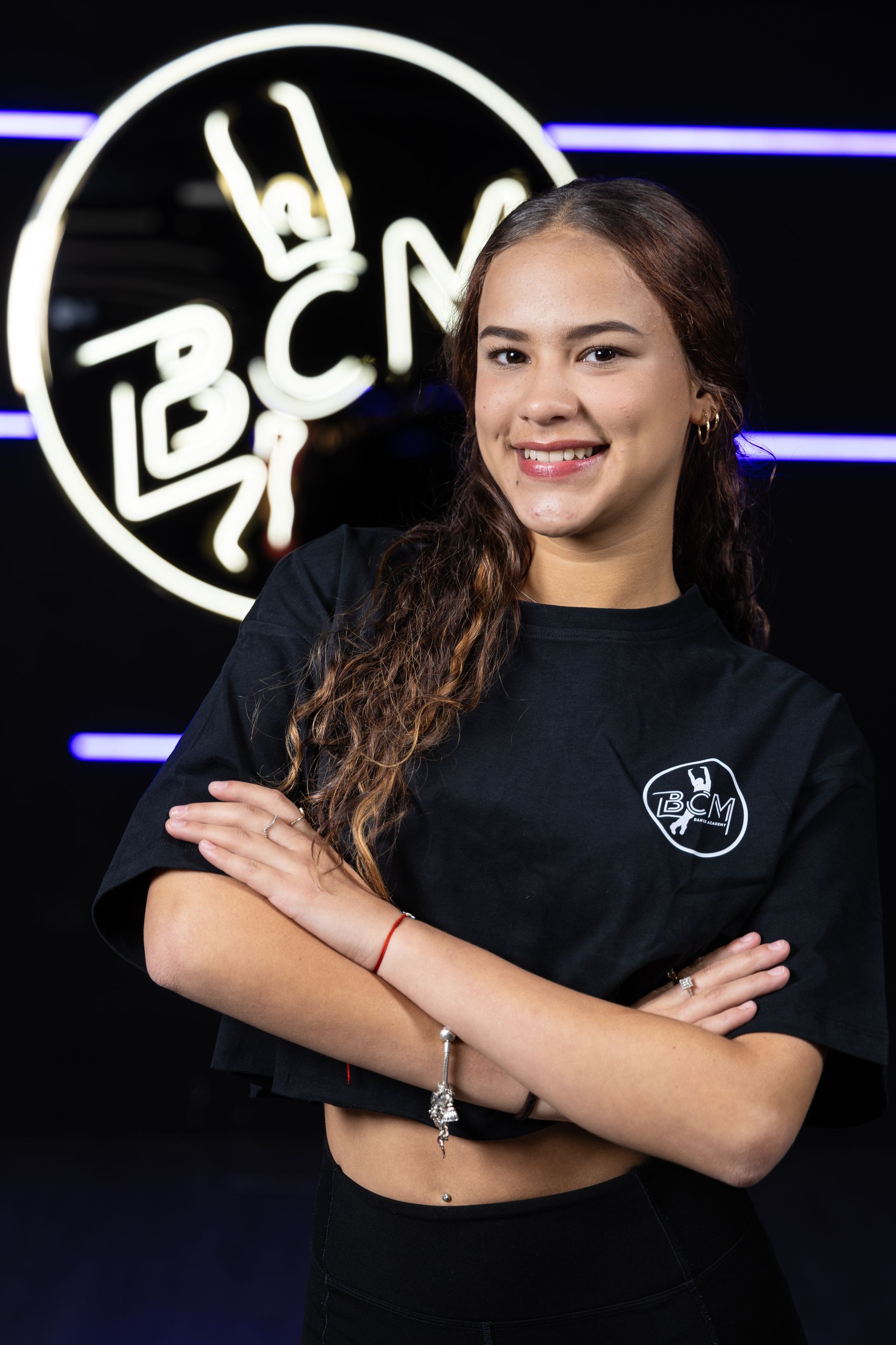 A young woman is posing for a picture with her arms crossed in front of a neon sign.