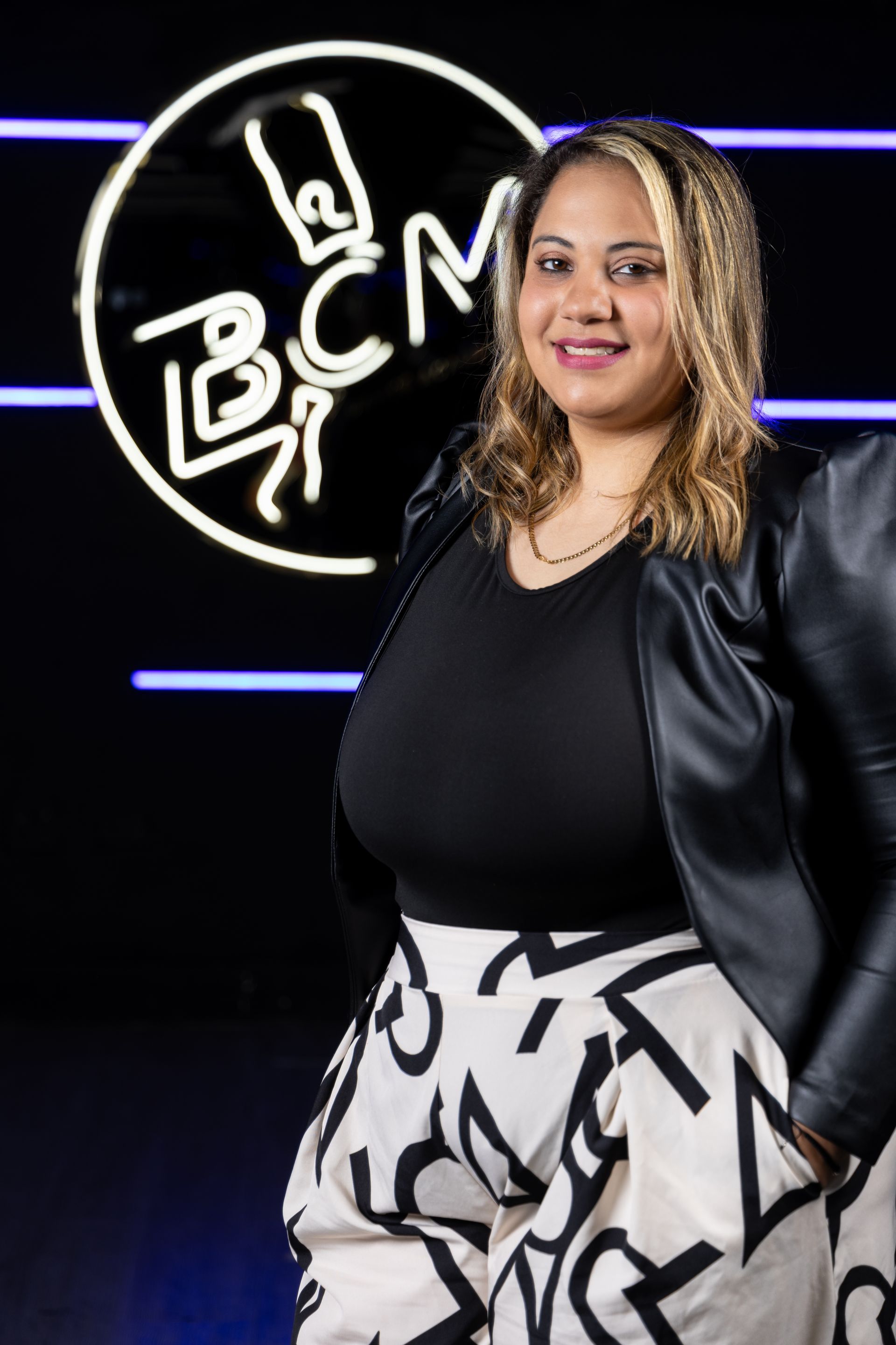 A woman in a black jacket and white skirt is standing in front of a neon sign.
