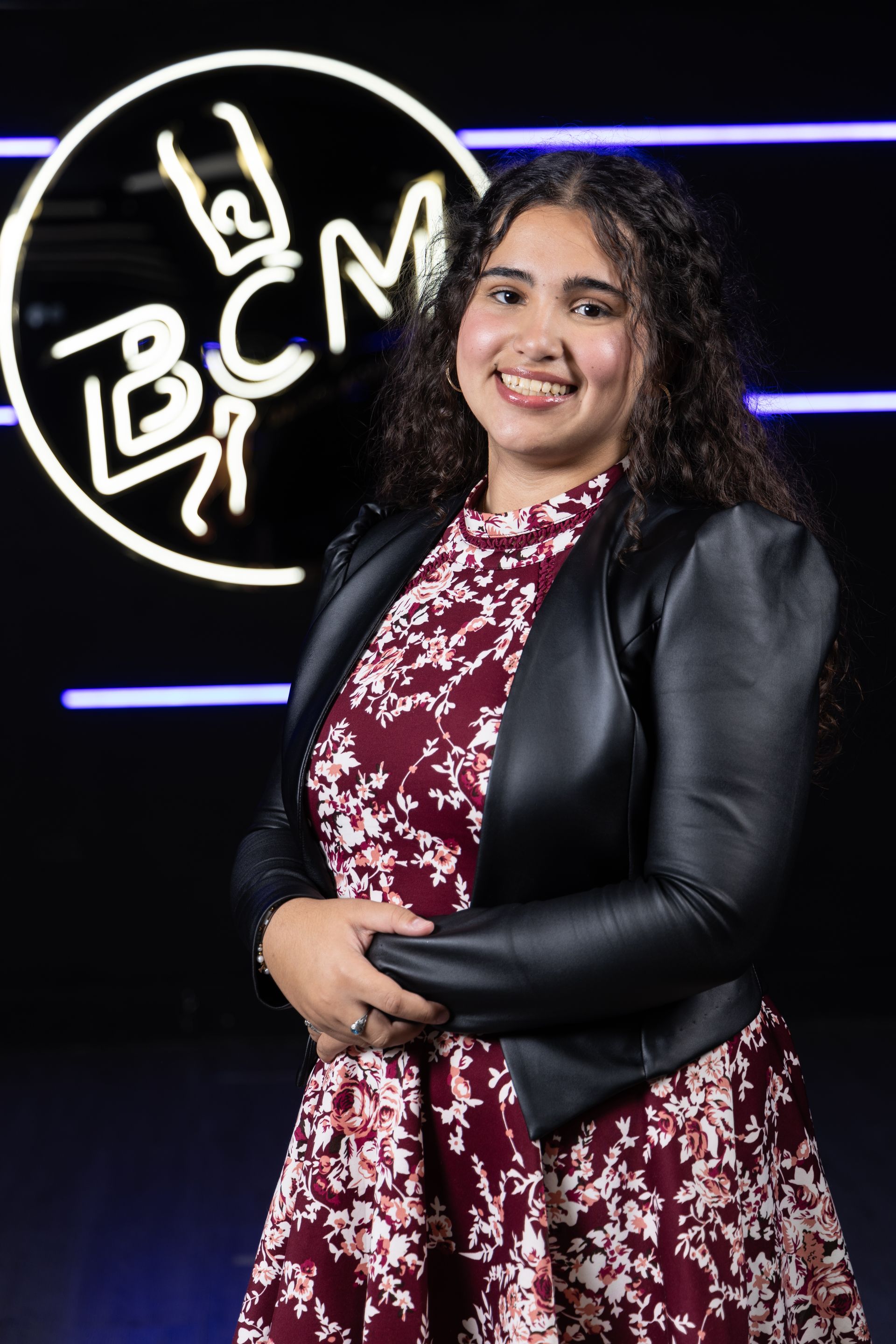 A woman in a floral dress and a black jacket is standing in front of a bcm logo.