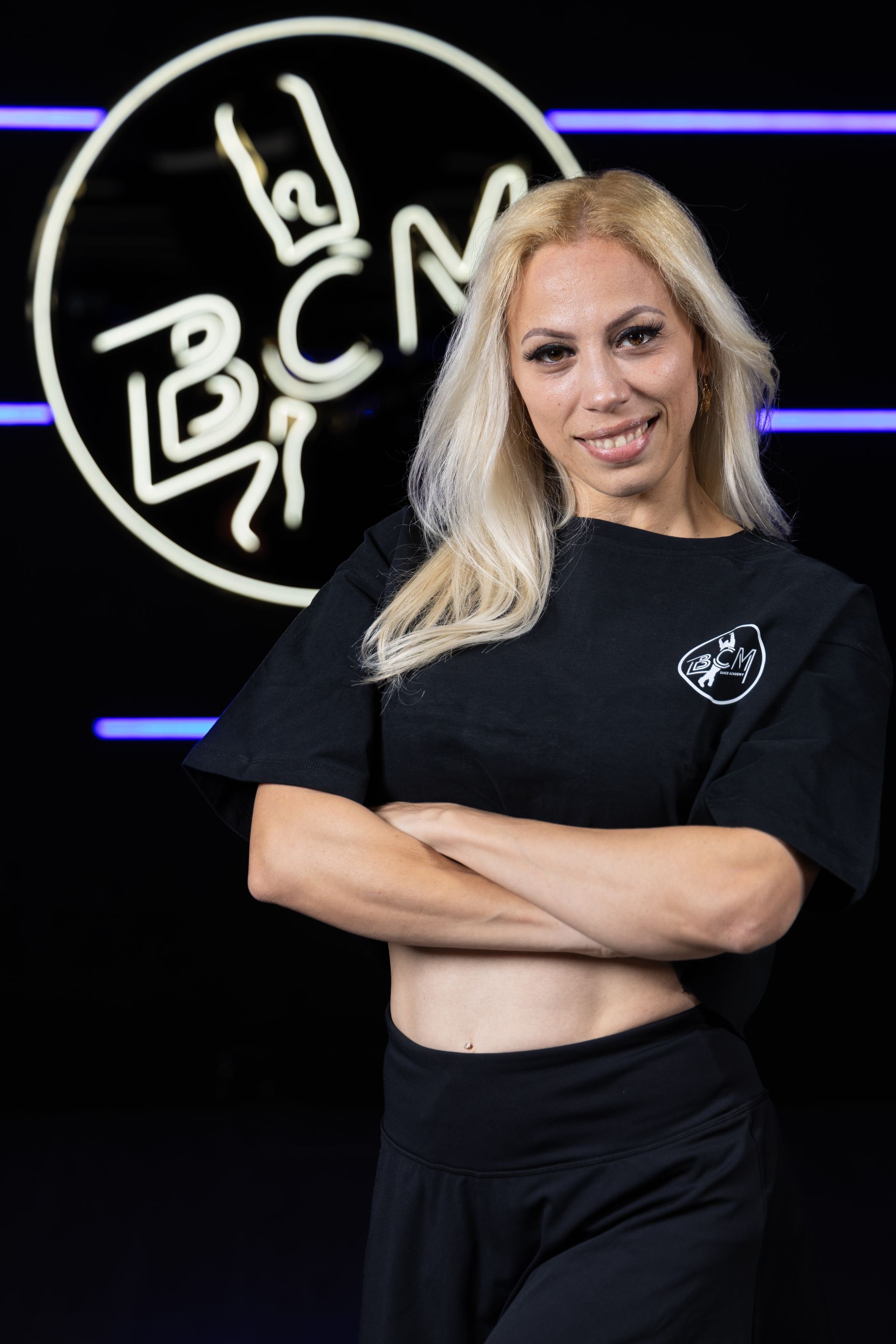 A woman is standing in front of a neon sign with her arms crossed.